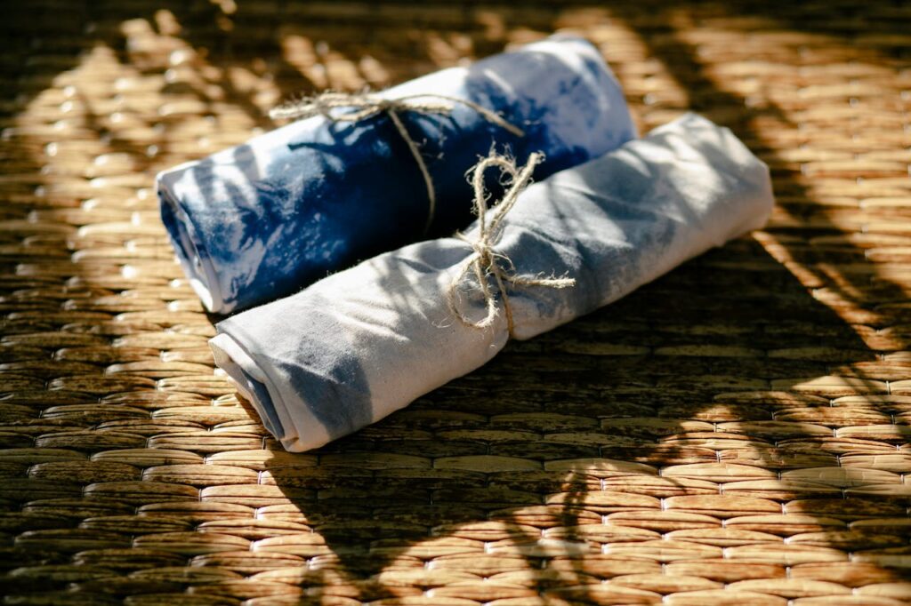 From above rolls of dyed white and blue textile tied with ropes placed on wicker surface in professional workshop with sunlight